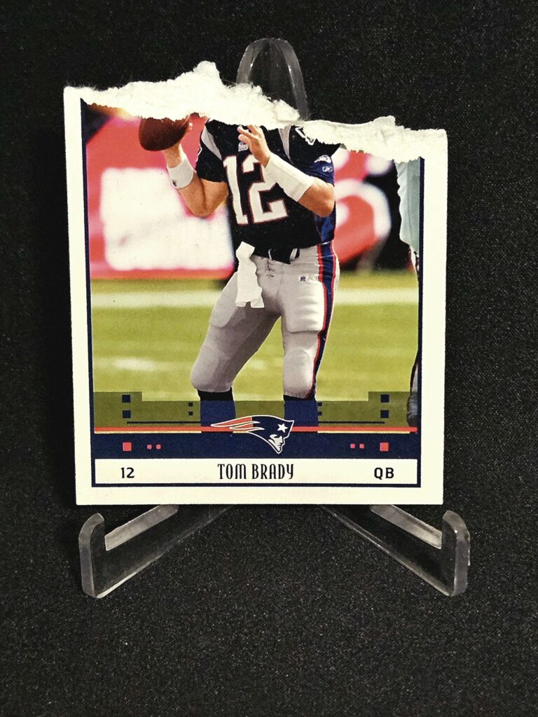 A picture of a 2005 Tom Brady Score football card is shown with a black background. The card is torn off at the top and is missing about 25% of its total stock. 